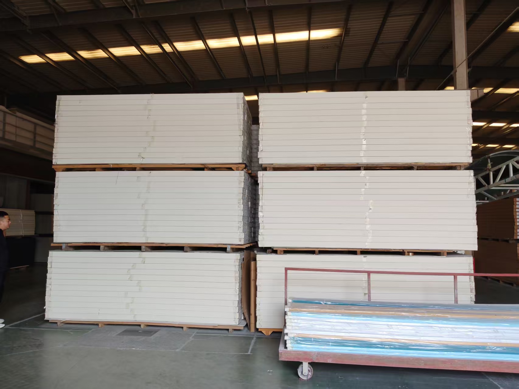 Interior of a finished-goods warehouse at a factory. A large number of white PVC foam boards are neatly stacked on wooden pallets in three layers; the smooth board surfaces are wrapped and secured with transparent plastic film. The ceiling is of steel structure with long strip LED lights. In the lower right corner, a wheeled red metal cart holds long profiles packed in blue-and-white transparent film. The floor is concrete; on the far left edge stands a man dressed in black.