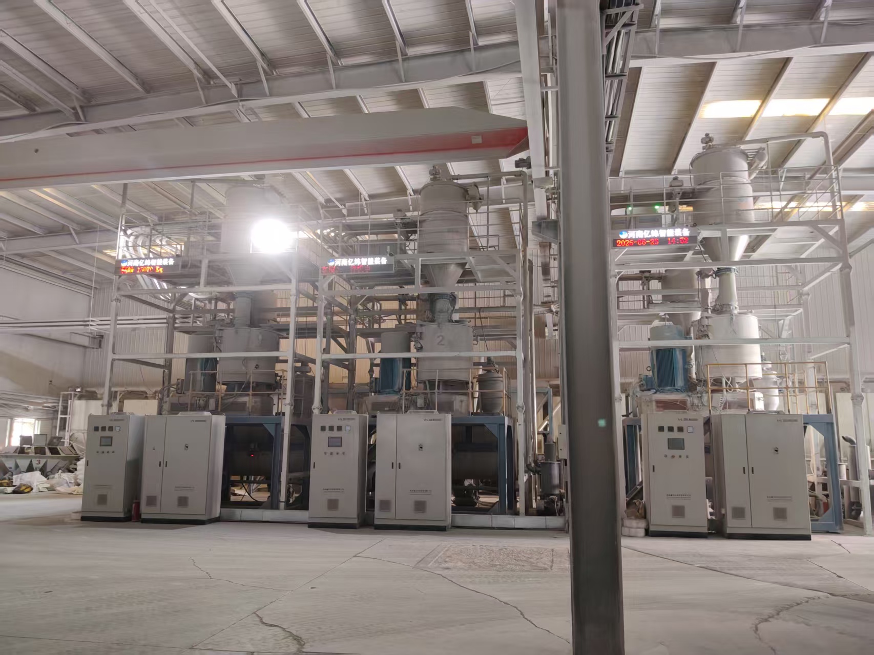 Interior of a large industrial production workshop showing a complete set of intelligent extrusion and batching equipment. The installation consists of multi-level steel work platforms, conical-top vertical silos, drive motors, and a row of grey electrical control cabinets below. Electronic display screens mounted on the steel frame show the text "Henan Yiwei Intelligent Equipment" along with date and time. The building has a steel roof and columns; above spans the red-and-white girder of an overhead bridge crane. The concrete floor shows visible cracks, and a thick grey column stands on the right. The space is brightly lit.