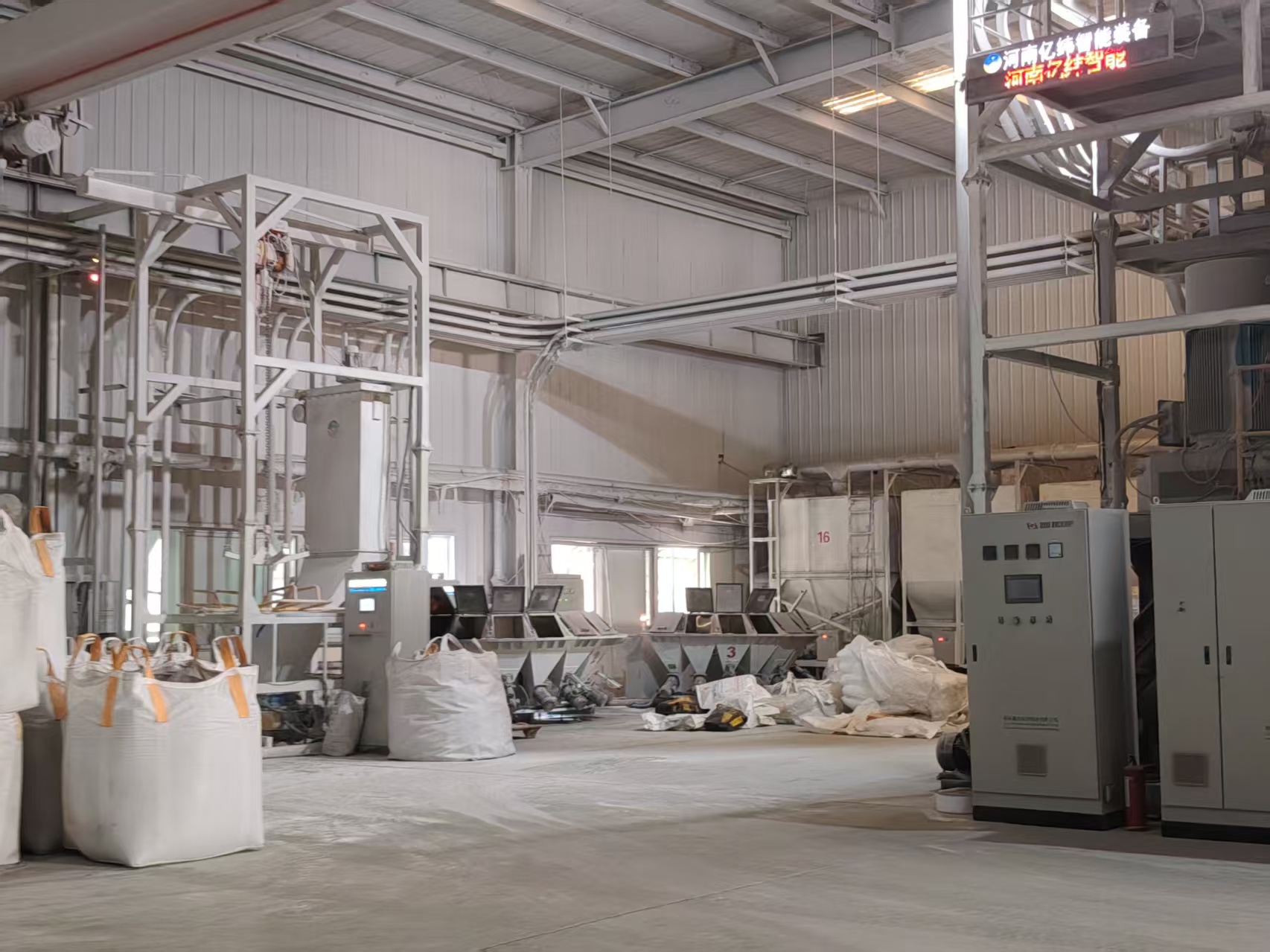 Interior of a raw-material batching workshop. In the left foreground sit several white bulk bags (FIBCs) with orange woven lifting straps. In the middle of the workshop are multiple sets of automatic batching hoppers (marked with the number "3"); behind them stands a large white storage silo marked "16", connected overhead by an extensive network of white conveying pipes and steel supports. On the right are grey electrical control cabinets and a red fire extinguisher. An electronic screen above displays "Henan Yiwei Intelligent Equipment". The walls are corrugated color steel panels, with natural light entering through side windows.
