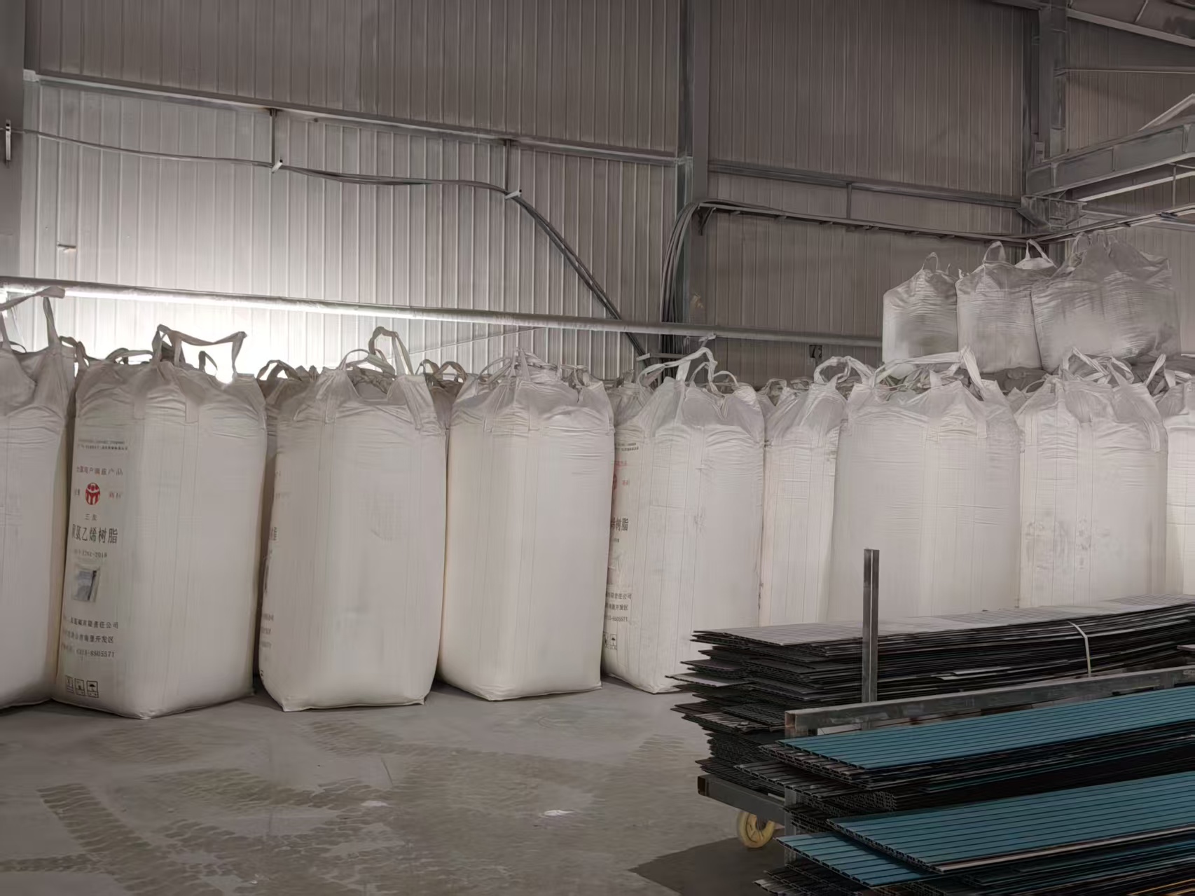 A corner of the raw-material storage area. A row of large white bulk bags stands neatly against a corrugated color-steel wall; one bag clearly shows the printed text "PVC resin (Polyvinyl Chloride Resin)" along with a brand logo and manufacturer information. In the lower right, a metal cart is stacked with black and blue PVC hollow profiles/grille panels. The floor is concrete with signs of material handling, and curved exposed cable conduits run along the wall.