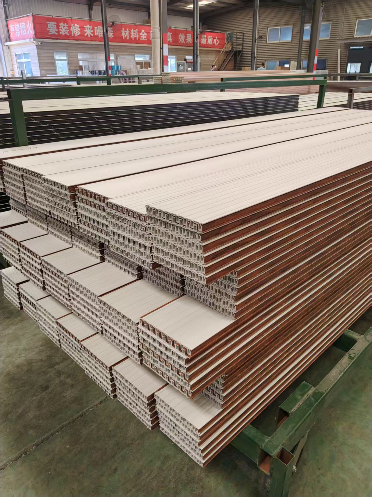 In the finished-goods warehouse racking area, large quantities of PVC decorative wall panels (hollow grille profiles) are neatly stacked on green metal racks. The profiles have a hollow square-cavity cross-section, with a white wood-grain surface and brown wood-imitating edge banding. Hanging on the rear wall are red banners reading "For renovation, come to Weisen — complete materials, authentic results, most reassuring" and "No open flames". The building features steel columns and glass windows; the concrete floor is clean and the stacks are dense and orderly.