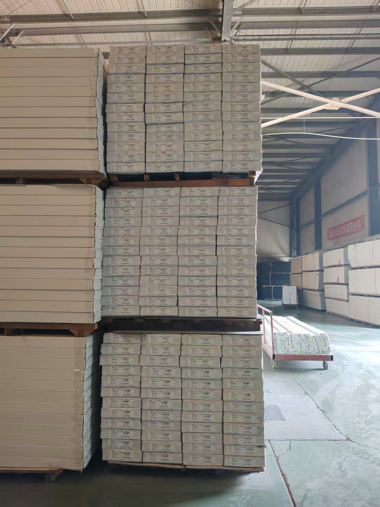 Interior of a finished-goods warehouse. Packaged PVC grille panels (labels printed with "PVC fluted panel" and specification codes) are neatly stacked in three layers on wooden pallets. In the distance on the right are more product stacks and a red cart carrying long items wrapped in blue packaging. A red slogan banner hangs on the rear wall; the roof is a steel skylight structure; the concrete floor is clean and the aisles are bright and spacious.